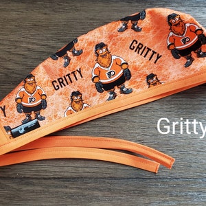 May include: Orange and black scrub hat with a repeated pattern of Gritty, the Philadelphia Flyers mascot, wearing a hockey jersey. The hat has a long orange tie.