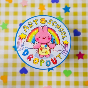 May include: A round, blue and white sticker with the text "ART SCHOOL DROPOUT" encircling a cartoon pink bunny holding a paintbrush. The bunny is in front of a rainbow and clouds. The background is a yellow and white checkered pattern with colorful star and heart-shaped decorations.