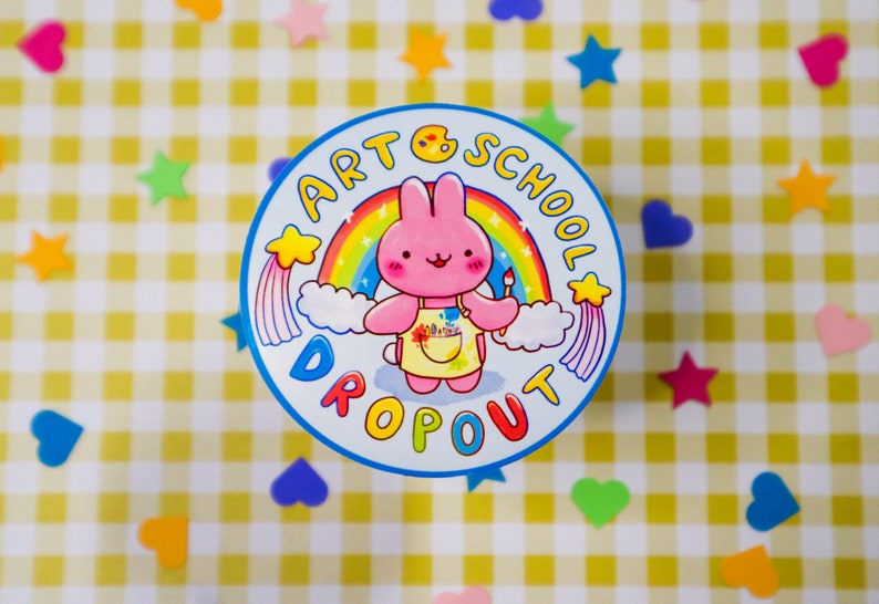 May include: A round, blue and white sticker with the text "ART SCHOOL DROPOUT" encircling a cartoon pink bunny holding a paintbrush. The bunny is in front of a rainbow and clouds. The background is a yellow and white checkered pattern with colorful star and heart-shaped decorations.