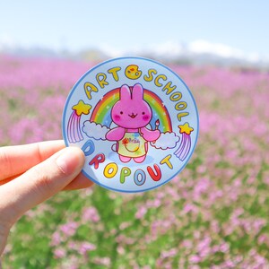 May include: A round sticker with a cartoon rabbit holding a paintbrush, wearing an apron, and standing in front of a rainbow. The sticker has the words "ART SCHOOL DROPOUT" in a circle around the rabbit.