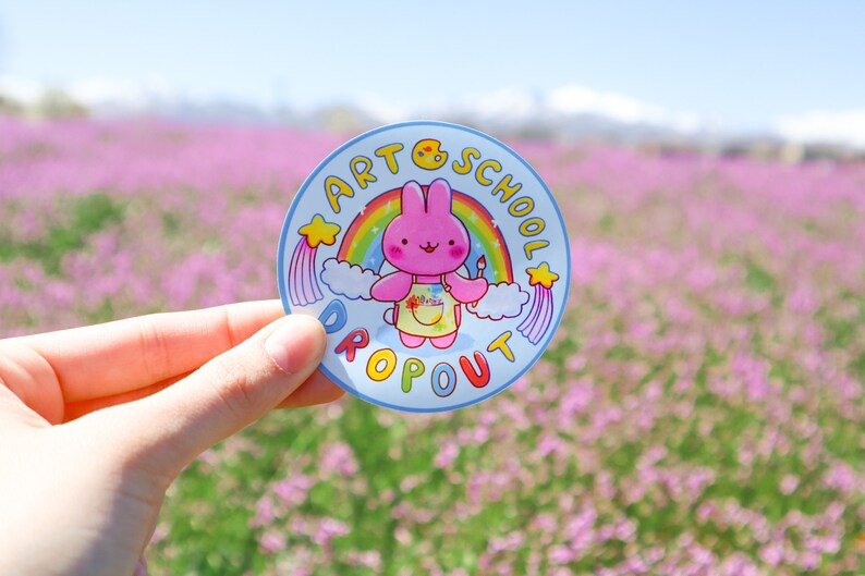May include: A round sticker with a cartoon rabbit holding a paintbrush, wearing an apron, and standing in front of a rainbow. The sticker has the words "ART SCHOOL DROPOUT" in a circle around the rabbit.