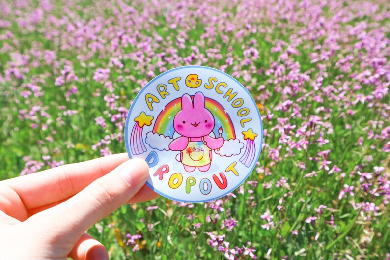 May include: A round sticker with a cartoon bunny wearing an apron and holding a paintbrush, with the text "ART SCHOOL DROPOUT" around a rainbow. The sticker is light blue with a white border and yellow stars.
