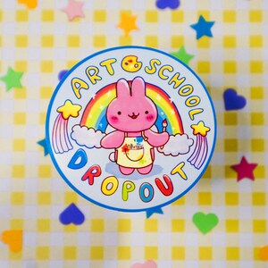 May include: A round sticker with a cartoon bunny wearing an apron and holding a paintbrush, set against a rainbow and clouds. The text "ART SCHOOL DROPOUT" encircles the image in blue. The background is a yellow and white checkered pattern with colorful star and heart-shaped decorations.
