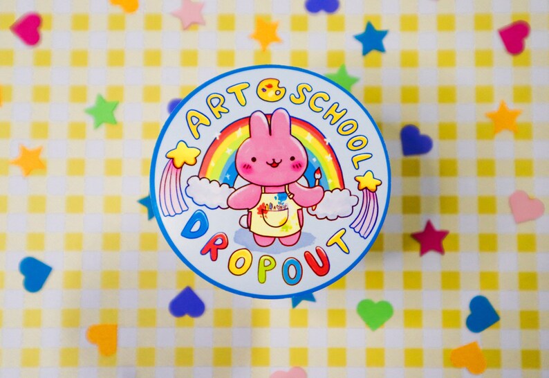 May include: A round sticker with a cartoon bunny wearing an apron and holding a paintbrush, set against a rainbow and clouds. The text "ART SCHOOL DROPOUT" encircles the image in blue. The background is a yellow and white checkered pattern with colorful star and heart-shaped decorations.