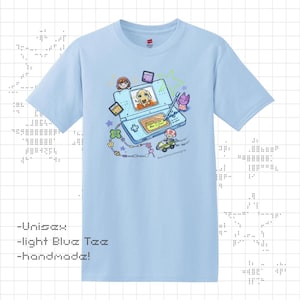 May include: Light blue unisex t-shirt with a colorful graphic of a handheld gaming device featuring a dog, characters, and the text "Best in Show". The shirt also has the text "-Unisex -light Blue Tee -handmade!"