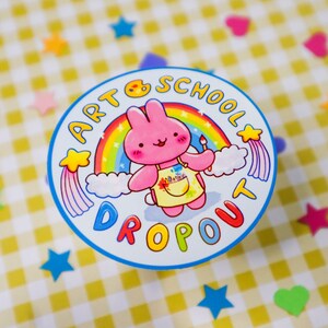 May include: A round sticker with a cartoon pink bunny wearing a yellow apron and holding a paintbrush. The sticker has the text "ART SCHOOL DROPOUT" around the image of the bunny and a rainbow. The background is a yellow and white checkered pattern.