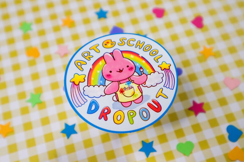 May include: A round sticker with a cartoon pink bunny wearing a yellow apron and holding a paintbrush. The sticker has the text "ART SCHOOL DROPOUT" around the image of the bunny and a rainbow. The background is a yellow and white checkered pattern.