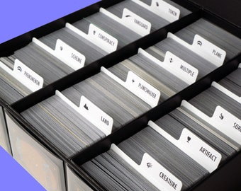 May include: A black card box with dividers for organizing trading cards. The dividers are labeled with different card types, including Creature, Artifact, Sorcery, Enchantment, Instant, Planeswalker, Land, Multiple, Plane, Conspiracy, Scheme, Vanguard, and Token. The text "CARD BOX AND CARDS NOT INCLUDED" is printed on the bottom right corner of the box.