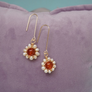 May include: Gold-tone earrings featuring a central orange-red bead surrounded by small white pearls. The earrings have a floral design and long, curved hooks. The earrings are displayed on a lavender pillow.