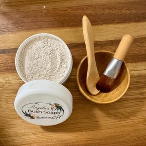 May include: A white container with a label that reads "Angela's Bush Soaps handmade with care" and a white powder inside. A wooden bowl with a wooden spoon and a small brush is next to the container.