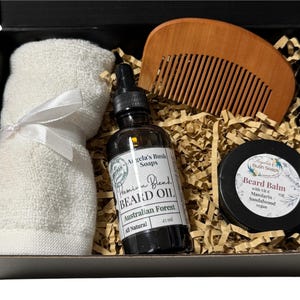 May include: A black gift box filled with beard care products. The box contains a wooden comb, a bottle of beard oil labelled "Premium Blend Beard Oil Australian Forest All Natural", a small jar of beard balm labelled "Beard Balm with Vit E Mandarin Sandalwood Vegan", and a white hand towel.