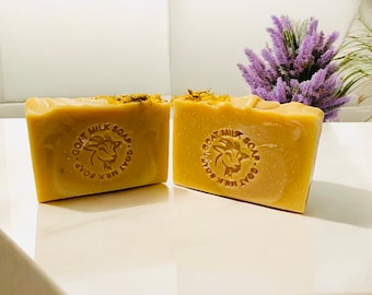 May include: Two rectangular bars of goat milk soap. The soap is a light tan colour with a stamped logo and text that reads "GOAT MILK SOAP". The top of the soap is decorated with dried flower petals. The soap is on a white surface.