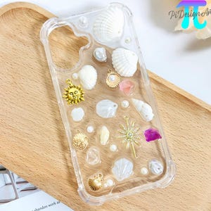 May include: Clear phone case decorated with seashells, pearls, and gold charms. The case features a sun, starburst, and seashell charms, along with a small piece of purple stone. The case is on a wooden tray.