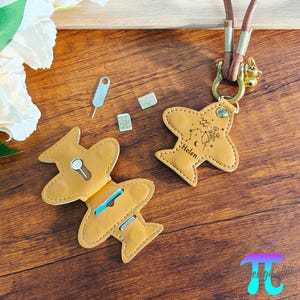 以下が含まれることがあります： タン色の革製飛行機型カードホルダー。鍵穴のデザインで、開くと青いカードが見えます。お揃いの飛行機型キーホルダーには「Helen」という名前と星座のデザインがあり、茶色のコードから吊り下げられています。SIMカードツールが含まれています。