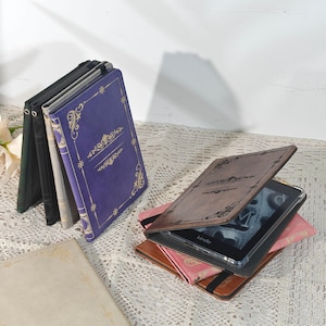 Może przedstawiać: Kolekcja etui na Kindle w stylu książkowym w różnych kolorach, w tym fioletowym, brązowym i czarnym. Okładki zdobią dekoracyjne, złote wzory filigranowe i są otwarte i zamknięte, prezentując urządzenia Kindle. Kindle jest widoczny w otwartych etui.