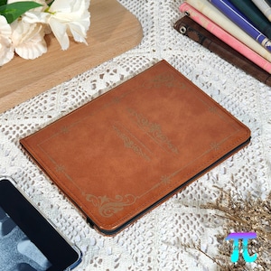 May include: A brown leather tablet case with decorative gold-colored filigree. The case is rectangular with a black zipper closure. The case is on a white lace surface with a wooden cutting board and flowers in the background.