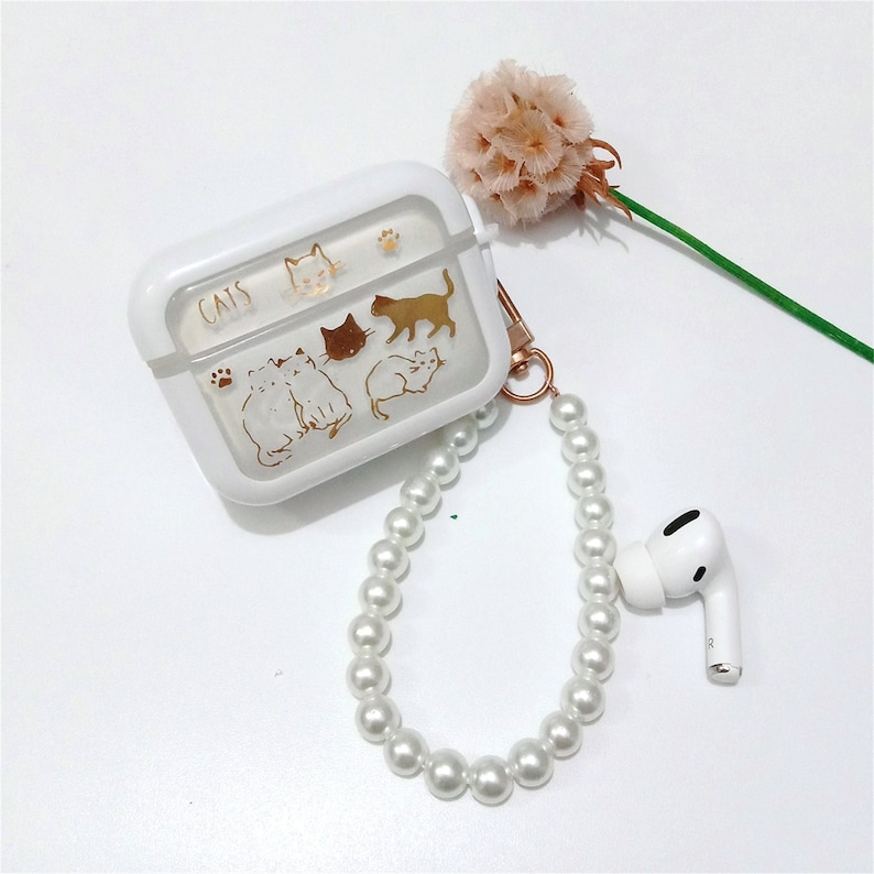 May include: A white phone case with a clear panel featuring a gold design of cats. The case has a gold chain with white pearl beads attached to it. The case also has the word "CATS" printed on it.
