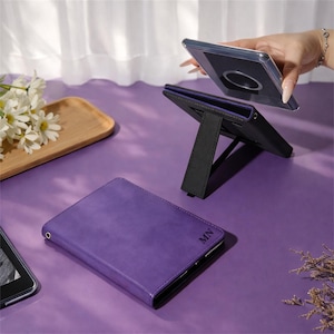 Puede incluir: Una funda para tableta de color morado con las iniciales MN grabadas en la cubierta frontal. La funda cuenta con un mecanismo de soporte plegable integrado que permite colocarla en posición vertical. Se ve una funda transparente con anillo magnético.