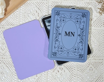 Könnte beinhalten: Eine hellviolette Tablethülle neben einer blauen Tablethülle mit den Initialen "MN" in einem dekorativen Oval. Die blaue Hülle hat ein verziertes Bordürendesign. Die Hüllen liegen auf einer weißen Spitzentischdecke.
