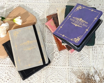 Puede incluir: Una colección de fundas para teléfono con forma de libro en varios colores, incluyendo gris, negro, morado y marrón. La funda morada presenta el texto "Gone with the Wind" en letras doradas. Cada funda tiene un diseño dorado decorativo.