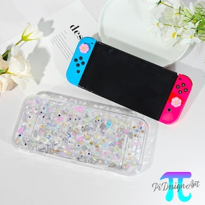 Può includere: Una console Nintendo Switch nera con controller blu e rosa, adornata da bottoni a forma di fiore. Sotto, una custodia trasparente piena di frammenti iridescenti colorati. Sulla custodia è stampata la parola "Carry".