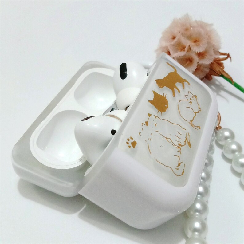 May include: White Airpods case with a gold design of cats. The case is open and the Airpods are inside.