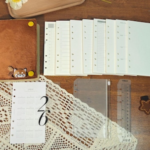 May include: A brown plush journal with a red panda design, accompanied by various planner inserts, a clear ruler, and a star-shaped bookmark. The planner inserts include lined, grid, and calendar pages. The calendar page shows the numbers "2" and "6".