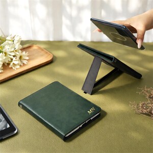Puede incluir: Una funda de teléfono de cuero verde oscuro con un monograma dorado "MN" se muestra junto a un soporte para teléfono negro. Una mano sostiene un teléfono inteligente sobre el soporte. La escena está ambientada sobre una superficie verde con flores y una bandeja de madera.