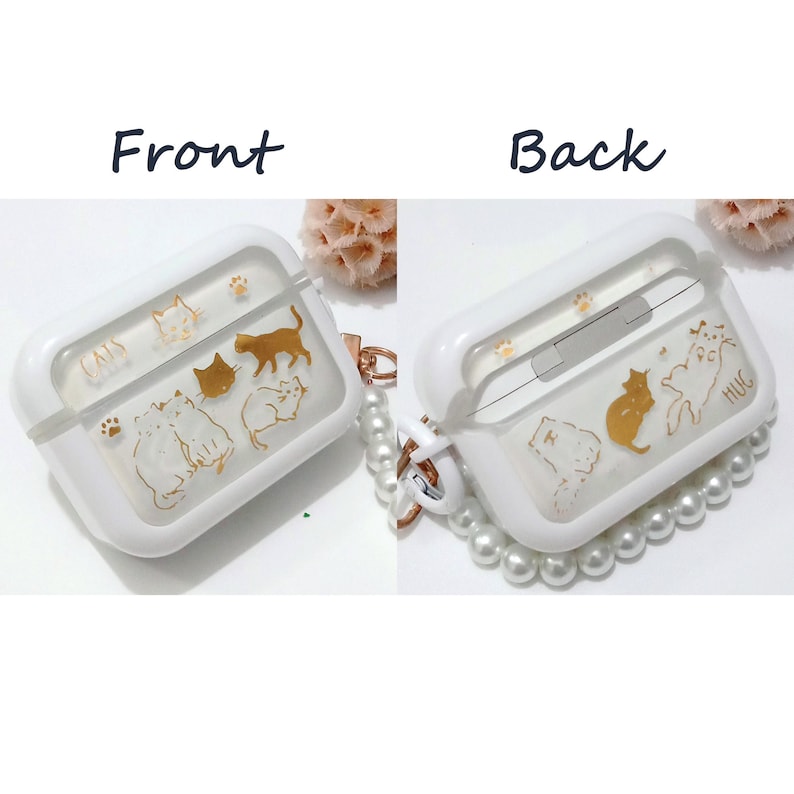 May include: White Airpods case with a clear back and a gold design of cats. The case has a pearl chain attached to it.