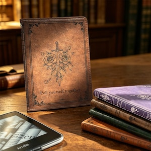 Puede incluir: Una funda para lector electrónico de textura de cuero marrón con un gráfico central de una espada entrelazada con dos rosas y el texto "Pull yourself together". Cerca hay una pila de cuatro libros de colores con motivos de rosas y un lector.
