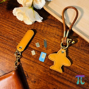 以下が含まれることがあります： 茶色の革製飛行機キーホルダー。茶色の革ストラップと金色の金具が付いています。キーホルダーには小さなベルが付いています。SIMカードとSIMカード取り出しツールも写っています。