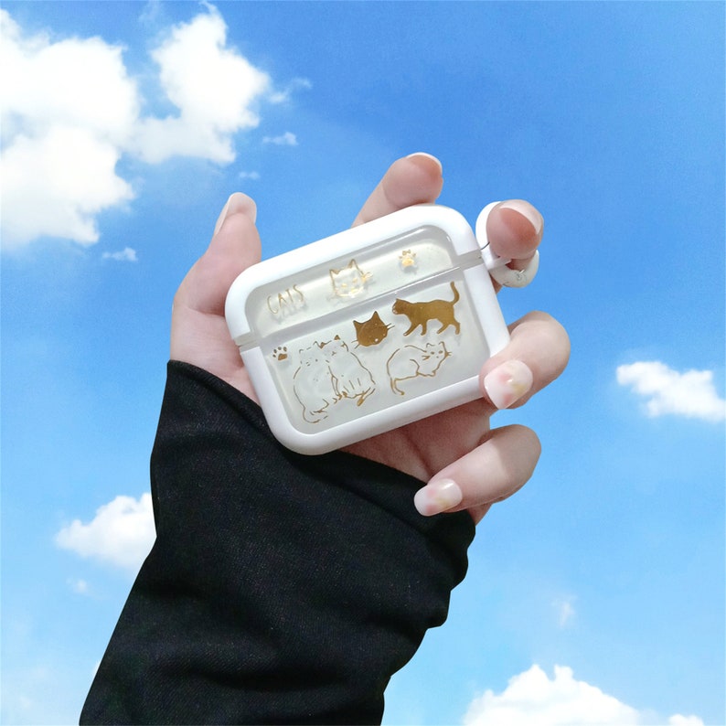 May include: A clear white phone case with a gold design of cats. The case has a cutout for the charging port and a small hole for the microphone. The case is being held by a hand.