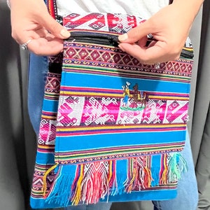 May include: A colorful crossbody bag with a vibrant woven design. The bag features a blue base with intricate patterns in pink, red, and white, and a fringed bottom. It has a black zipper and a striped strap. The bag is approximately 12 inches wide and 14 inches tall.