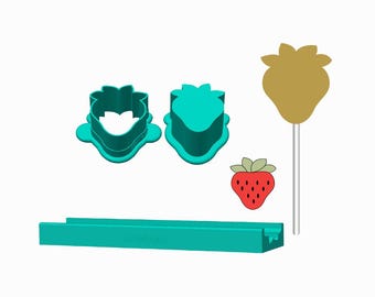 May include: A set of two teal strawberry-shaped cookie cutters, a long rectangular teal base, a gold strawberry-shaped topper on a white stick, and a small red strawberry illustration with green leaves and black seeds on a white background.