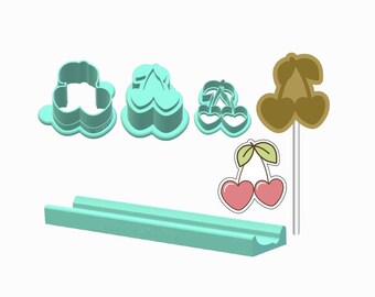 Peut inclure: Ensemble de découpoirs à biscuits sur le thème des cerises et une sucette cerise. Les découpoirs sont bleu clair et présentent divers motifs de cerises. La sucette est marron avec un bâton blanc. Un objet rectangulaire bleu clair est également présent.