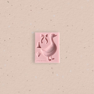 Puede incluir: Un molde rectangular de silicona rosa con formas hundidas de un pato, un lazo, un gorro de fiesta y una pata palmeada. El texto LITTLE BIKKIE está grabado en la esquina inferior izquierda del molde, diseñado para crear figuras moldeadas.