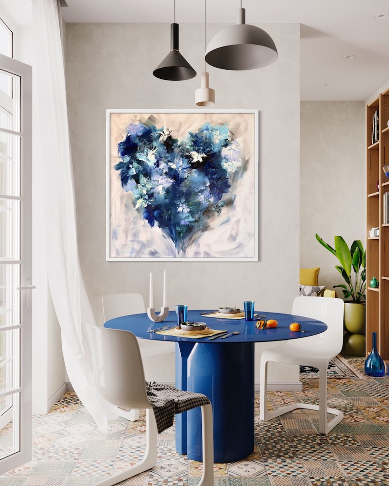 May include: A blue and white abstract painting of a heart shape, framed in white, hanging on a wall in a dining room with a blue table and white chairs.