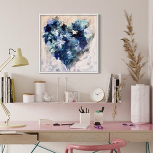 May include: A framed abstract painting of a blue heart shape with white and light blue accents. The painting is on a white wall above a desk with a pink surface.