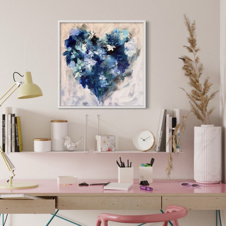 May include: A framed abstract painting of a blue heart shape with white and light blue accents. The painting is on a white wall above a desk with a pink surface.