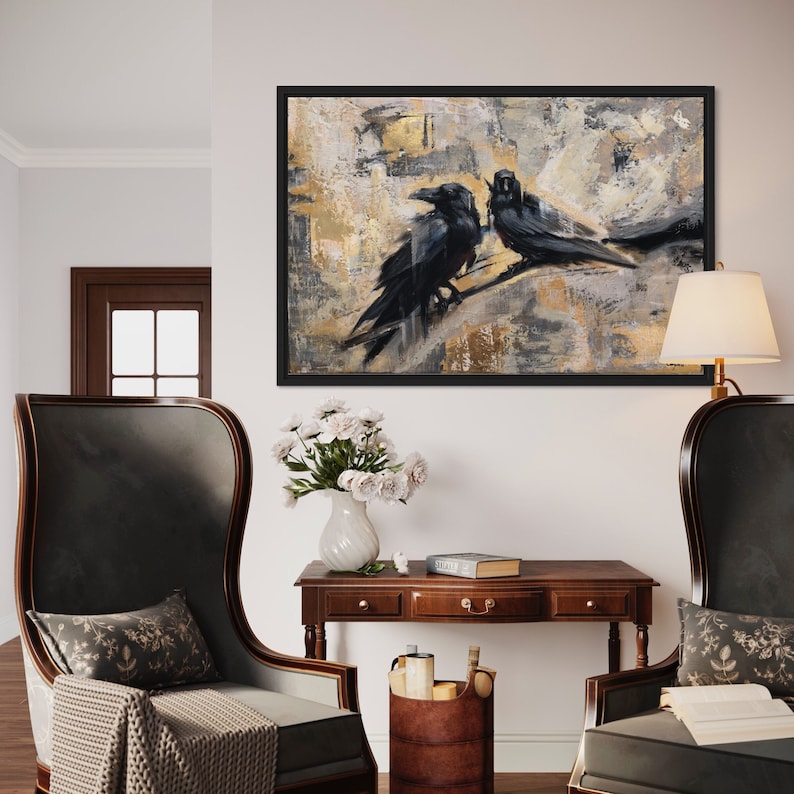 May include: Two black ravens perched on a branch in a painting with a gold and gray abstract background. The painting is framed in black and hanging on a white wall. The wall is decorated with two chairs, a table, a vase with white flowers, and a book. The chairs are upholstered in dark gray fabric and have a high back. The table is made of dark wood and has a drawer. The vase is made of white ceramic and is filled with white flowers. The book is brown and has a white spine.