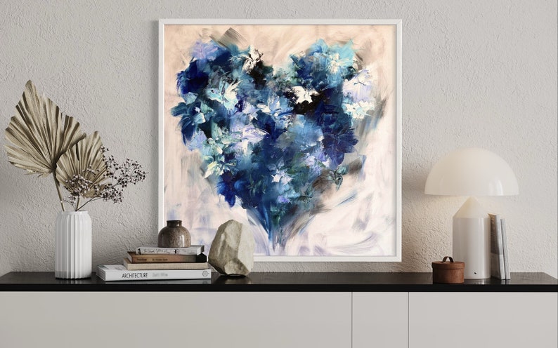 May include: Abstract painting of a blue heart-shaped floral arrangement on a white background. The painting is framed in a white frame and is hanging on a white wall.