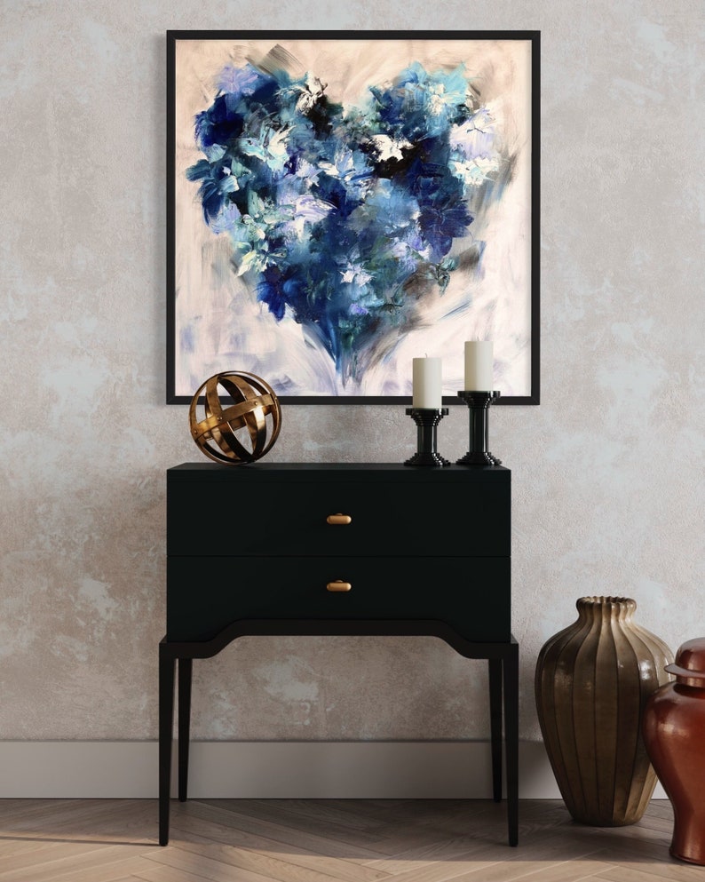 May include: A black framed painting of a blue and white abstract heart-shaped floral design hangs above a black two-drawer dresser with gold knobs. A gold metal sphere sits on the dresser, and two candlesticks with white candles are on either side of the sphere.