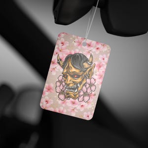 May include: A rectangular air freshener with a Japanese demon illustration. The design features a tan background with pink cherry blossoms. The demon has yellow skin, black hair, red eyes, and horns. A white string is attached to the top.