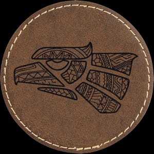 May include: A round, brown leather coaster with a stitched edge. The coaster features a detailed black graphic of an eagle's head with intricate tribal patterns. The design is centered on the coaster.