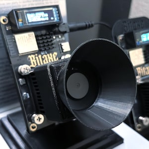 May include: A black electronic device with a screen displaying text, connected to a black funnel-shaped component. The device has the word "Bitaxe" printed on it. The device is mounted on a black stand.