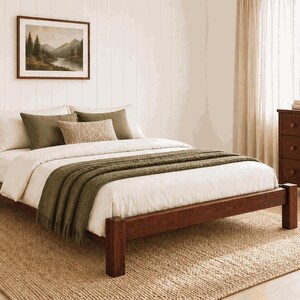 May include: A wooden bed frame with a white comforter, olive green throw blanket, and pillows. A framed landscape painting hangs above the bed. The bed sits on a woven jute rug. A dark wooden dresser is to the right.