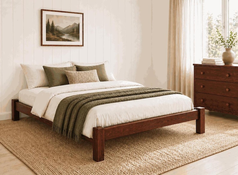 May include: A wooden bed frame with a white comforter, olive green throw blanket, and pillows. A framed landscape painting hangs above the bed. The bed sits on a woven jute rug. A dark wooden dresser is to the right.
