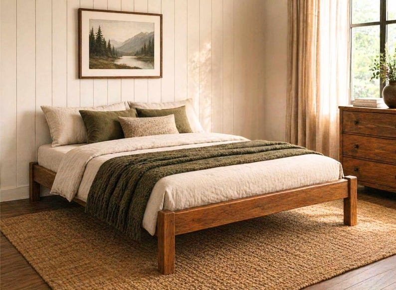 May include: A wooden bed frame with a neutral-toned comforter and olive green throw blanket. The bed is on a woven jute rug. A framed landscape painting hangs above the bed, and a wooden dresser is visible in the background.