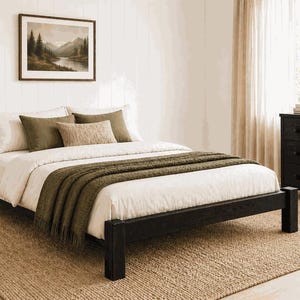 May include: A bedroom scene featuring a black wooden bed frame with white bedding and olive green throw blanket and pillows. A framed landscape painting hangs above the bed. A woven rug and a black dresser complete the room.