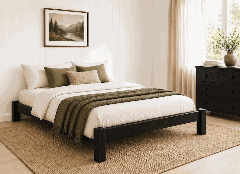May include: A bedroom scene featuring a black wooden bed frame with white bedding and olive green throw blanket and pillows. A framed landscape painting hangs above the bed. A woven rug and a black dresser complete the room.